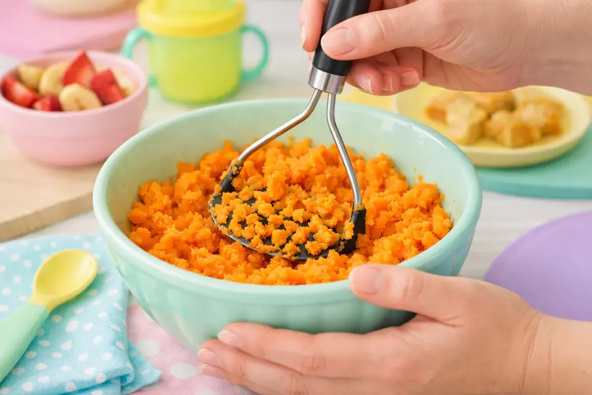 Step 1: Bright orange cooked sweet potato being mashed with a potato masher in a pastel mint bowl
